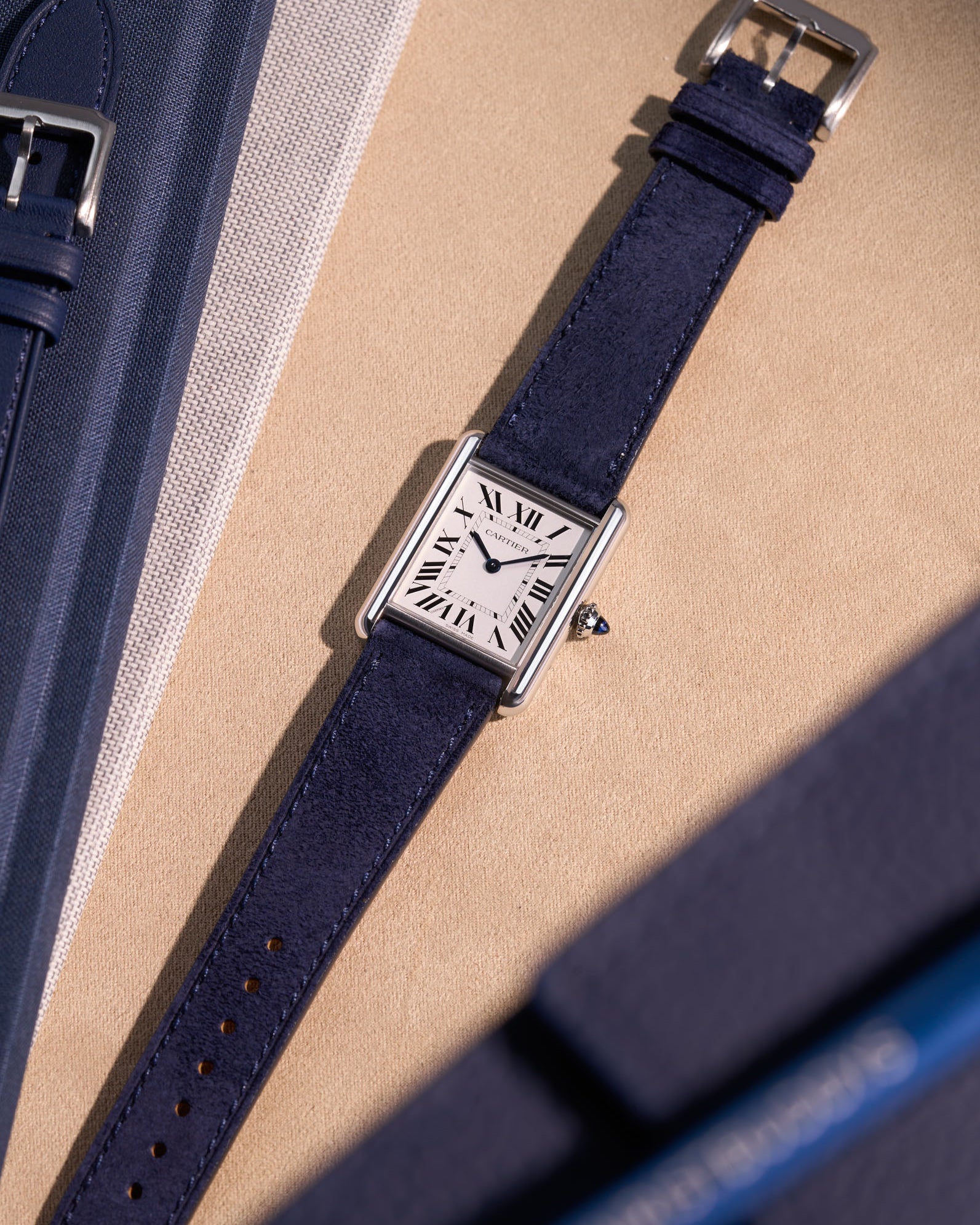 Cartier Tank Must Large with Dark Blue Suede leather strap, flat lay on textured beige surface with clasp visible