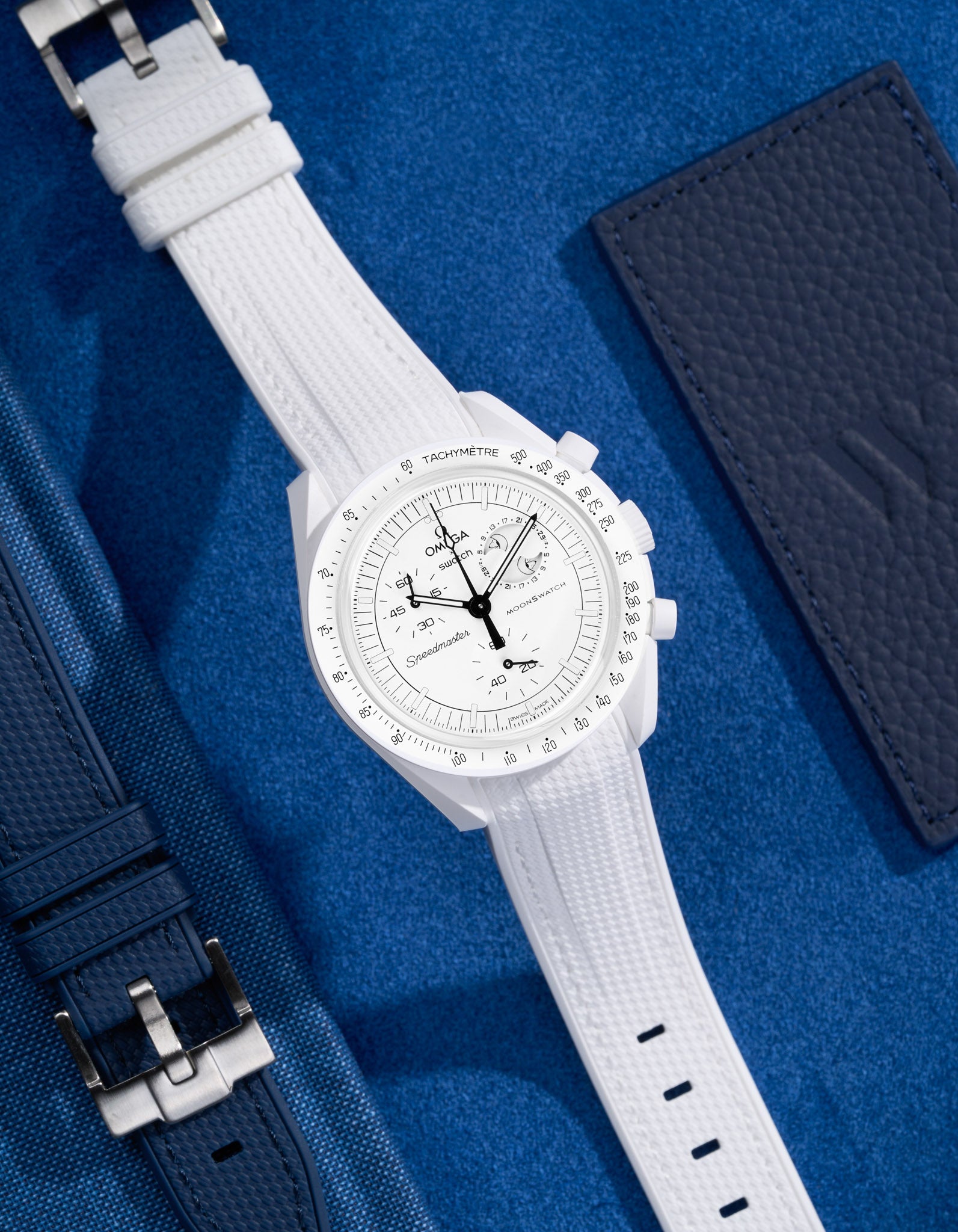 Flat lay of MoonSwatch Mission to the Moonphase with white vertech strap and accessories in background