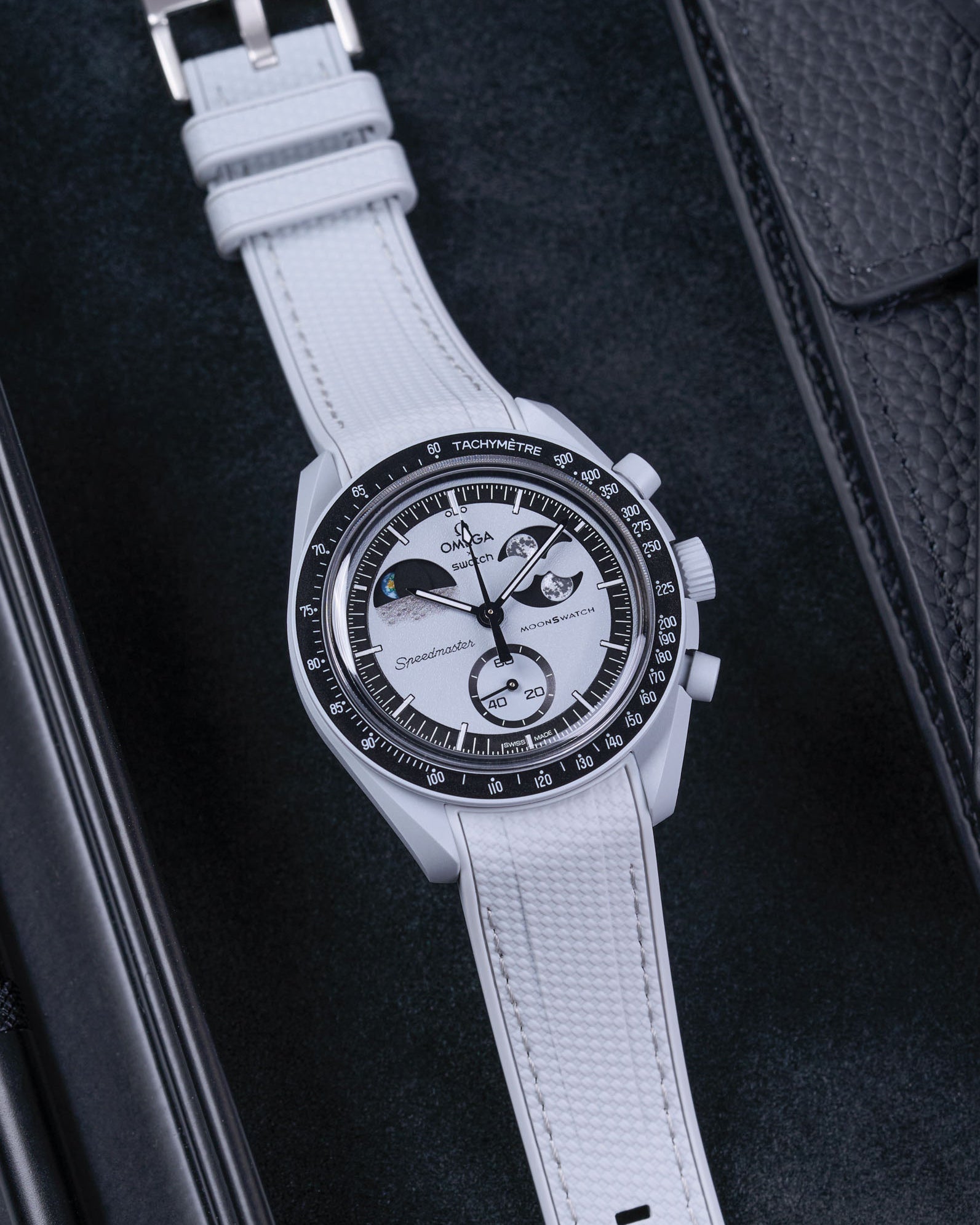 Flat lay of MoonSwatch Mission to EarthPhase with grey rubber strap, watch facing up with clasp visible on a table with black background