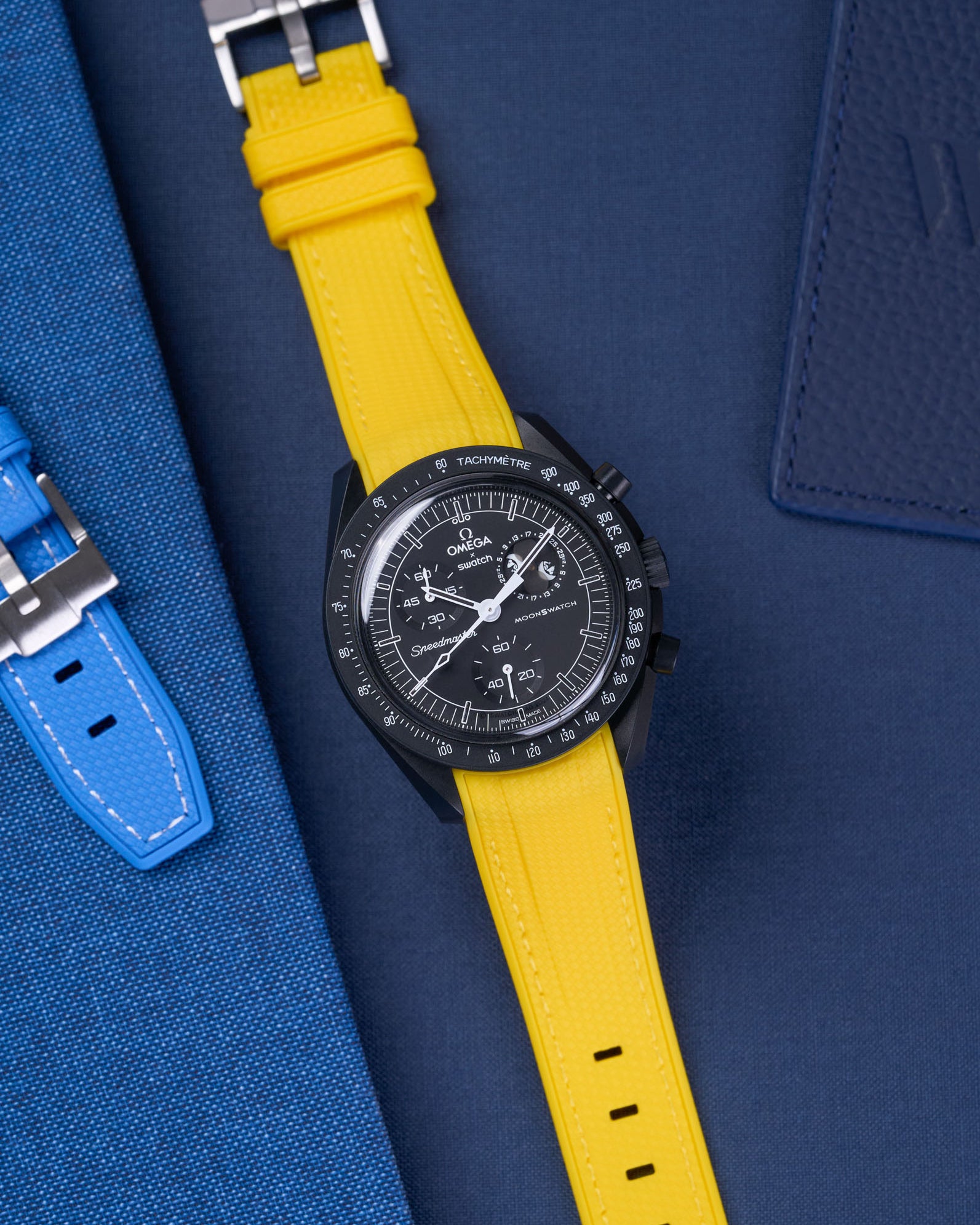 MoonSwatch Mission to Moonphase new Moon with vertech yellow rubber strap placed on blue fabric surface next to blue pouch