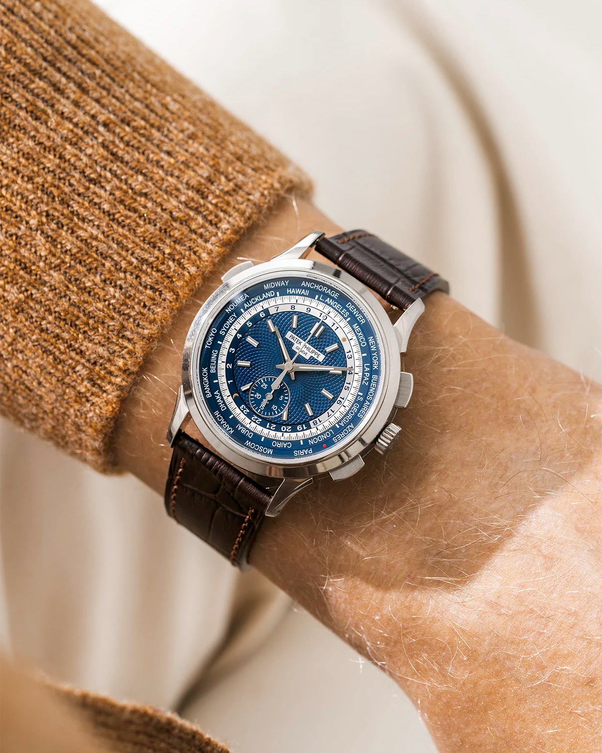 Patek Worldtimer watch with a blue face and dark brown wristbuddys alligator watch strap on a wrist, against a neutral background