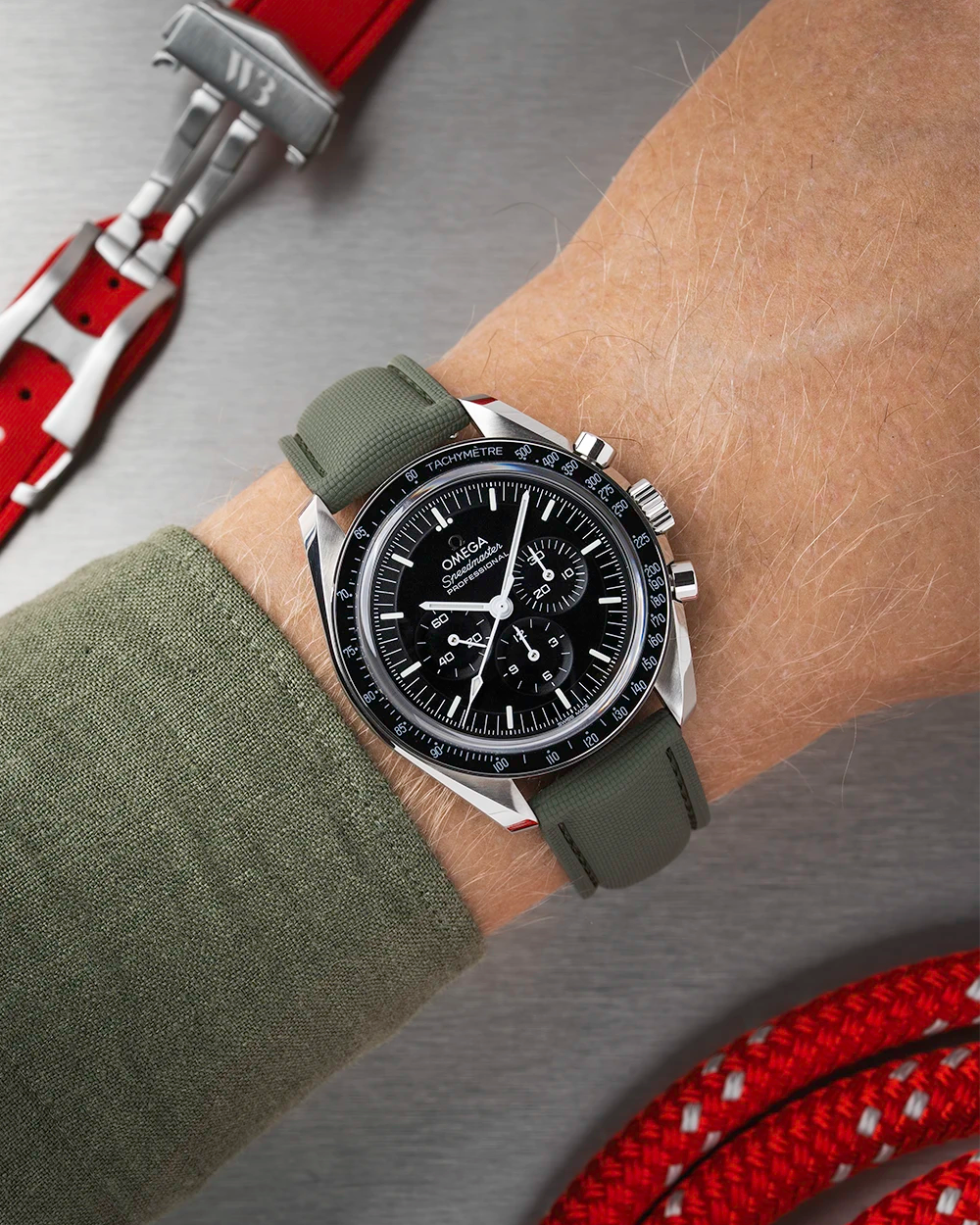 Wrist wearing a black Omega Speedmaster  with a detailed face on a wrist with a green sleeve.
