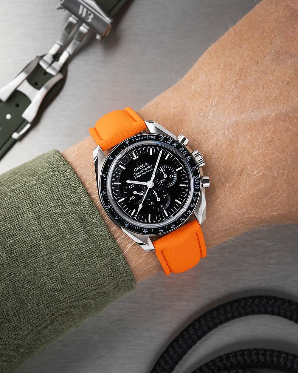 Wrist wearing a black Omega Speedmaster with a orange strap  with a detailed face on a wrist with a green sleeve.