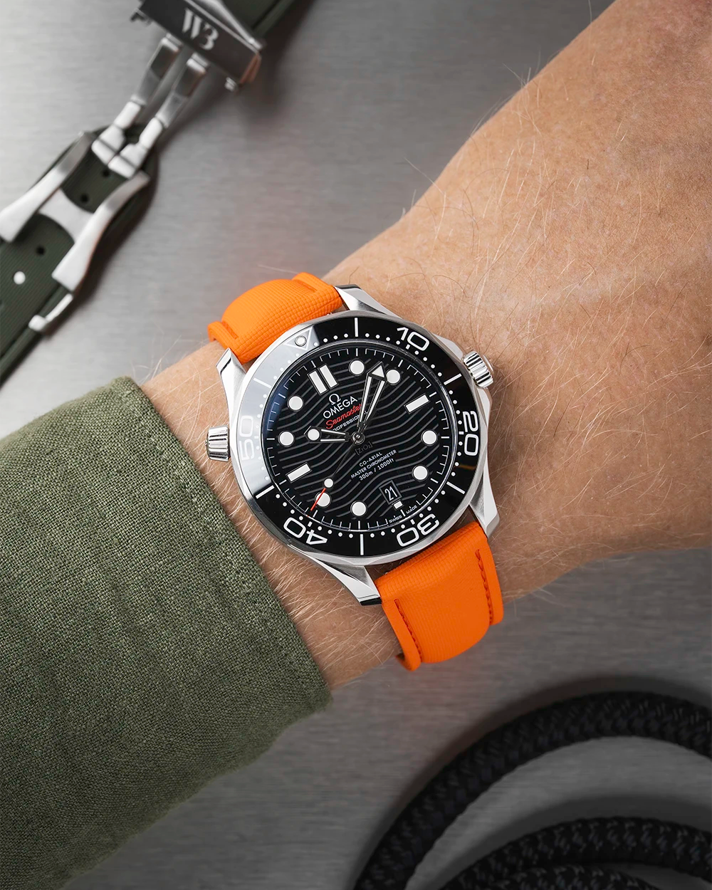 Wrist wearing a black Omega Seamaster with a orange strap  with a detailed face on a wrist with a green sleeve.