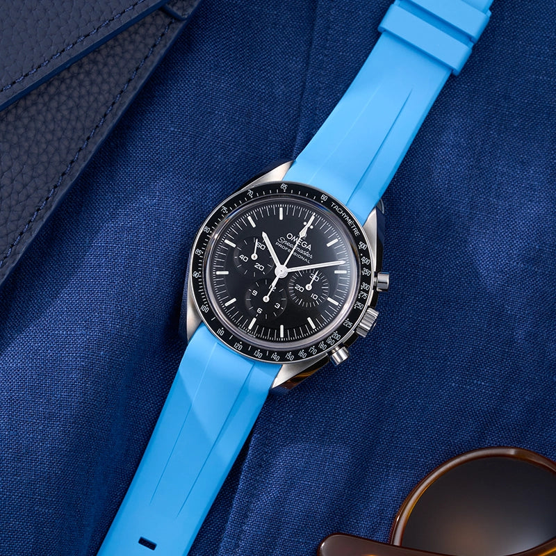 Omega Speedmaster Professional with light blue FKM rubber strap placed on blue fabric with sunglasses nearby