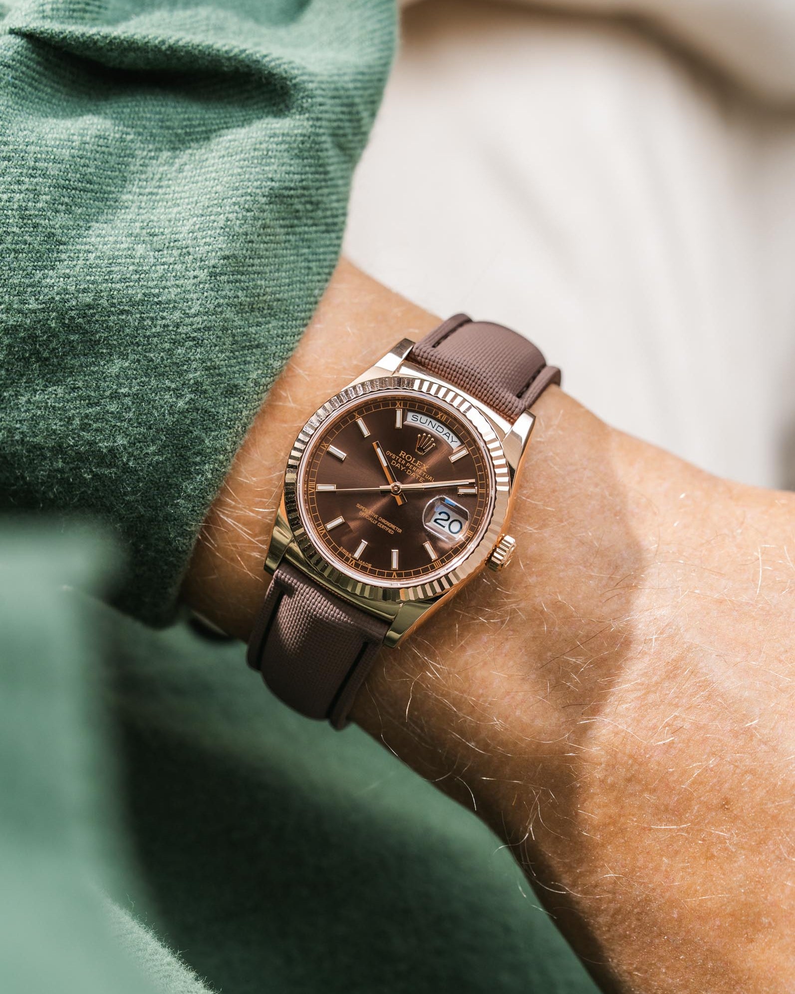 Brown Rolex DayDate in rose gold with leather strap on a wrist, blurred green and beige background
