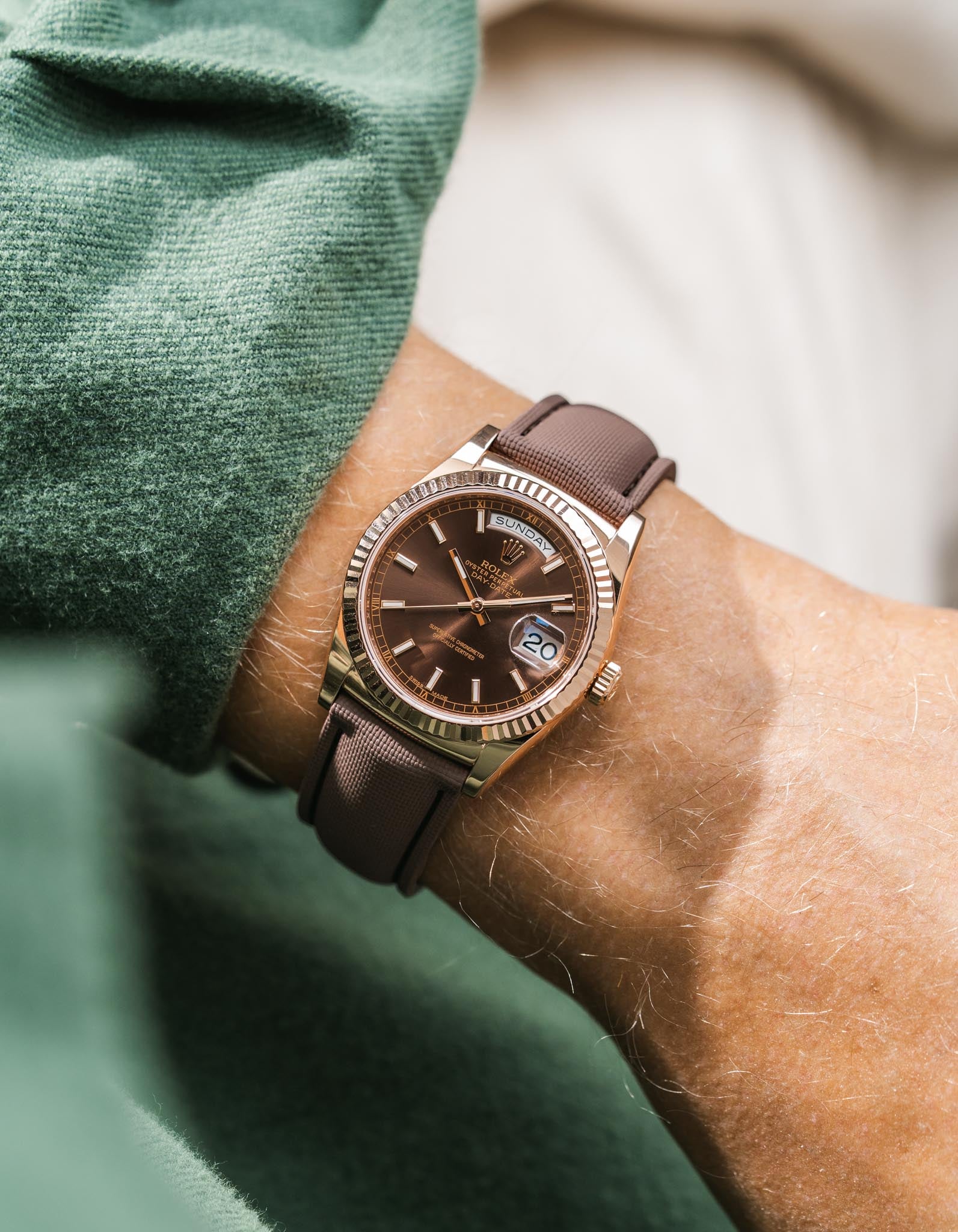 Brown Rolex DayDate in rose gold with leather strap on a wrist, blurred green and beige background