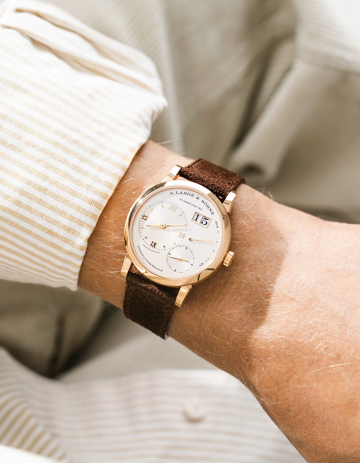 A Lange Shöne in rose gold with a brown suede leather strap from Wristbuddys on a wrist, wearing a beige sleeve.