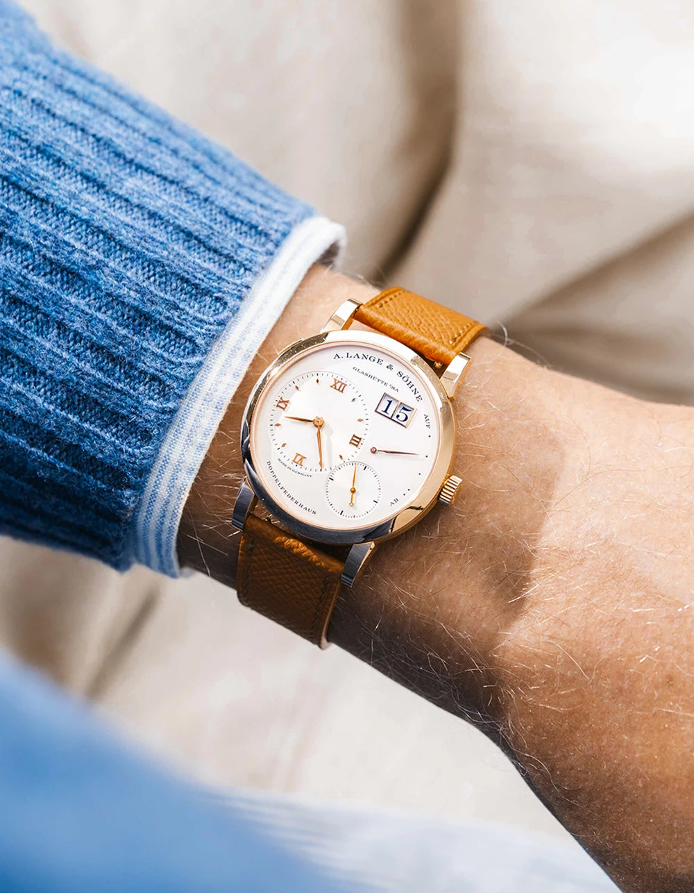 A Lange Shöne in rose gold with a orange epsom leather watch strap from Wristbuddys on a wrist, wearing a blue sleeve.