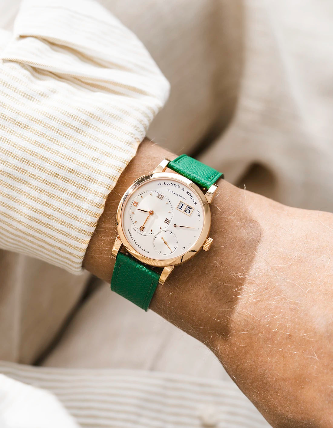 A Lange Shöne in rose gold with a green epsom leather watch strap from Wristbuddys on a wrist, wearing a beige sleeve.