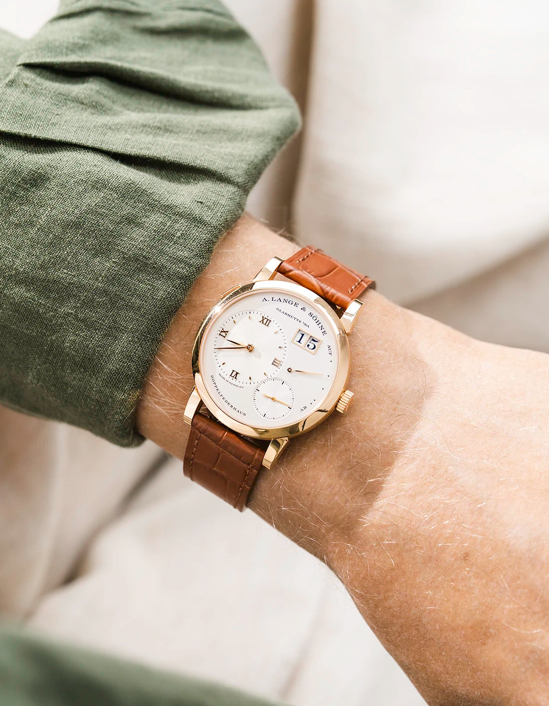 A Lange Shöne in rose gold with a brown embossed alligator leather strap from Wristbuddys on a wrist, wearing a green sleeve.