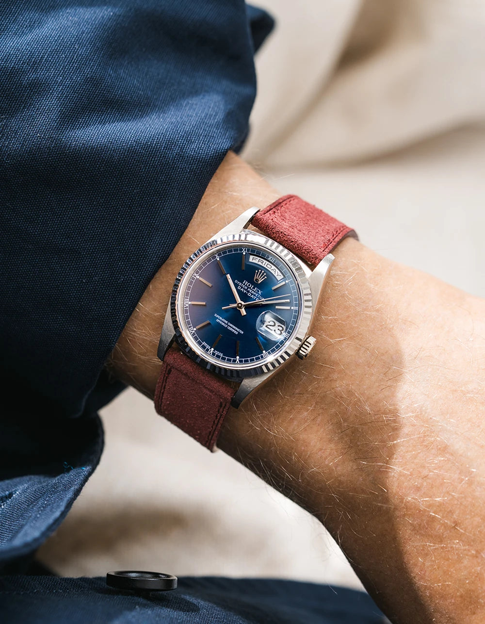 A rolex DayDate in white gold with a burgundy suede leather strap from Wristbuddys on a wrist, wearing a blue sleeve.