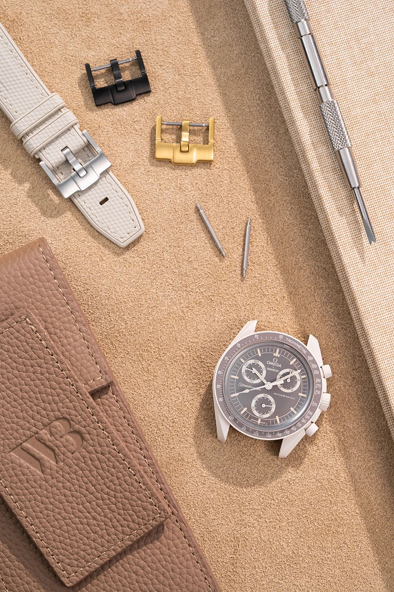 Wristbuddys watch accessories including MoonSwatch, leather pouch, interchangeable buckles and beige rubber strap placed on suede surface.