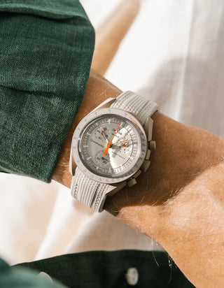 Beige rubber strap with cross-hatched texture on Omega x Swatch MoonSwatch Mission to Jupiter, wrist shot against white linen background, silver-grey dial with orange chronograph hands, cream bioceramic case, dark green jacket visible at left