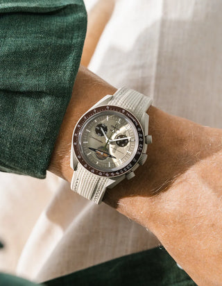 Beige rubber strap with fine cross-hatched ribbed texture on Omega x Swatch MoonSwatch Mission to Saturn, wrist shot against white linen fabric background, pale sage green dial with dark brown subdials and dark brown tachymeter bezel, light grey bioceramic case, dark green linen jacket visible at upper and lower left
