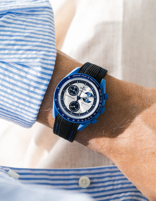 Black rubber strap with blue stitching fitted to an OMEGA x Swatch MoonSwatch Mission to the Super Blue Moonphase watch, worn on the wrist in a lifestyle shot with clothing visible