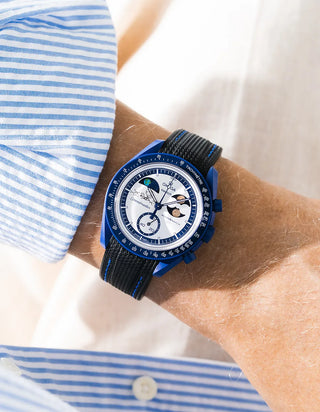 Black rubber strap with blue contrast stitching and diagonal ribbed texture on Omega x Swatch MoonSwatch Mission to Earthphase Moonshine Gold, wrist shot against cream fabric background, white dial with blue moonphase complication, blue and white striped shirt with white button visible at left and lower right