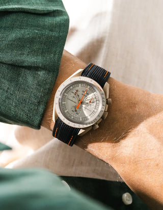 Black rubber strap with orange contrast stitching and diagonal ribbed texture on Omega x Swatch MoonSwatch Mission to Jupiter, wrist shot against white linen background, silver-grey dial with orange chronograph hands and silver tachymeter bezel, cream bioceramic case, dark green linen jacket with white button visible at left