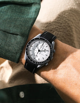 Black rubber strap with white contrast stitching and fine cross-hatched ribbed texture on Omega x Swatch MoonSwatch Mission to the Moon 1965, wrist shot against beige sandstone rock background, white dial with black subdials and black tachymeter bezel, white bioceramic case, dark green linen jacket with white button visible at lower left