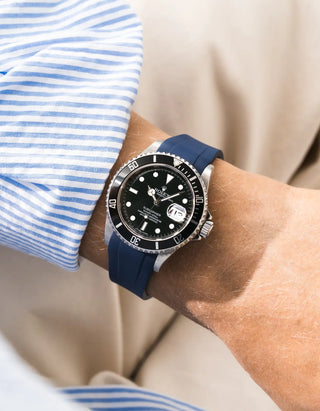 Blue FKM rubber strap integrated for original clasp on Rolex Submariner 16610LN full steel, black ceramic bezel, black dial, angled top-down wrist shot, smooth navy blue strap texture, light blue striped linen shirt sleeve, beige background