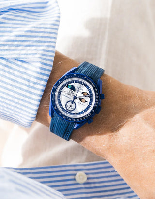 Blue rubber strap with diagonal ribbed texture on Omega x Swatch MoonSwatch Mission to Earthphase Moonshine Gold, wrist shot against cream fabric background, white dial with blue moonphase complication, blue and white striped shirt with white button visible at left and lower right