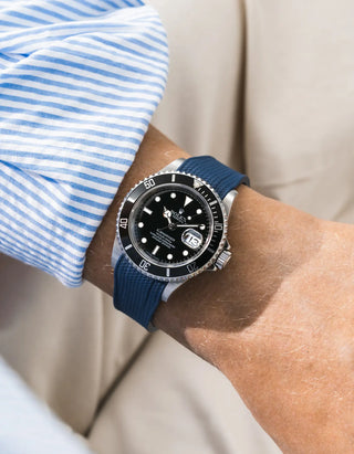 Blue Vertech FKM rubber strap integrated for original clasp on Rolex Submariner 16610LN full steel, black ceramic bezel, black dial, angled top-down wrist shot, Vertech crosshatch texture clearly visible on navy blue strap, light blue striped linen shirt sleeve, cream background