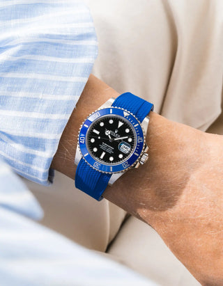 Blue Vertech FKM rubber strap integrated for original clasp on Rolex Submariner 126619LB "Cookie Monster" 18k white gold, blue ceramic bezel, black dial, angled top-down wrist shot, Vertech crosshatch texture clearly visible on royal blue strap matching bezel color, light blue striped linen shirt sleeve, cream background