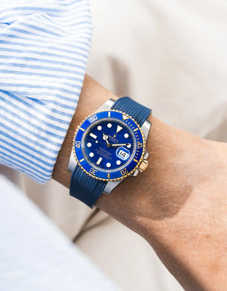 Blue Vertech FKM rubber strap on Rolex Submariner 116610LB Bluesy two-tone steel and yellow gold, blue ceramic bezel, blue dial, worn on wrist with blue striped shirt cuff, close-up front angle, white background