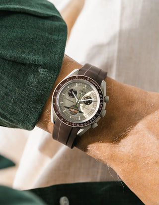Smooth plain dark brown rubber strap without texture on Omega x Swatch MoonSwatch Mission to Saturn, wrist shot against white linen fabric background, pale champagne-sage dial with dark brown subdials and dark brown tachymeter bezel, light grey bioceramic case, dark green linen jacket visible at upper left, shot from above
