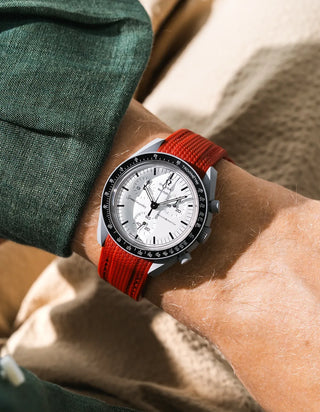 Burgundy red rubber strap with diagonal ribbed texture on Omega x Swatch MoonSwatch Mission to the Moon 1965, wrist shot against beige linen cushion background, white dial with black subdials and black tachymeter bezel, white bioceramic case, dark green linen jacket visible at upper left