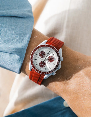 Burgundy rubber strap with diagonal ribbed texture on Omega x Swatch MoonSwatch Mission to Pluto, wrist shot against cream fabric background, white dial with burgundy subdials and burgundy tachymeter bezel, light grey bioceramic case, blue chambray shirt visible at left
