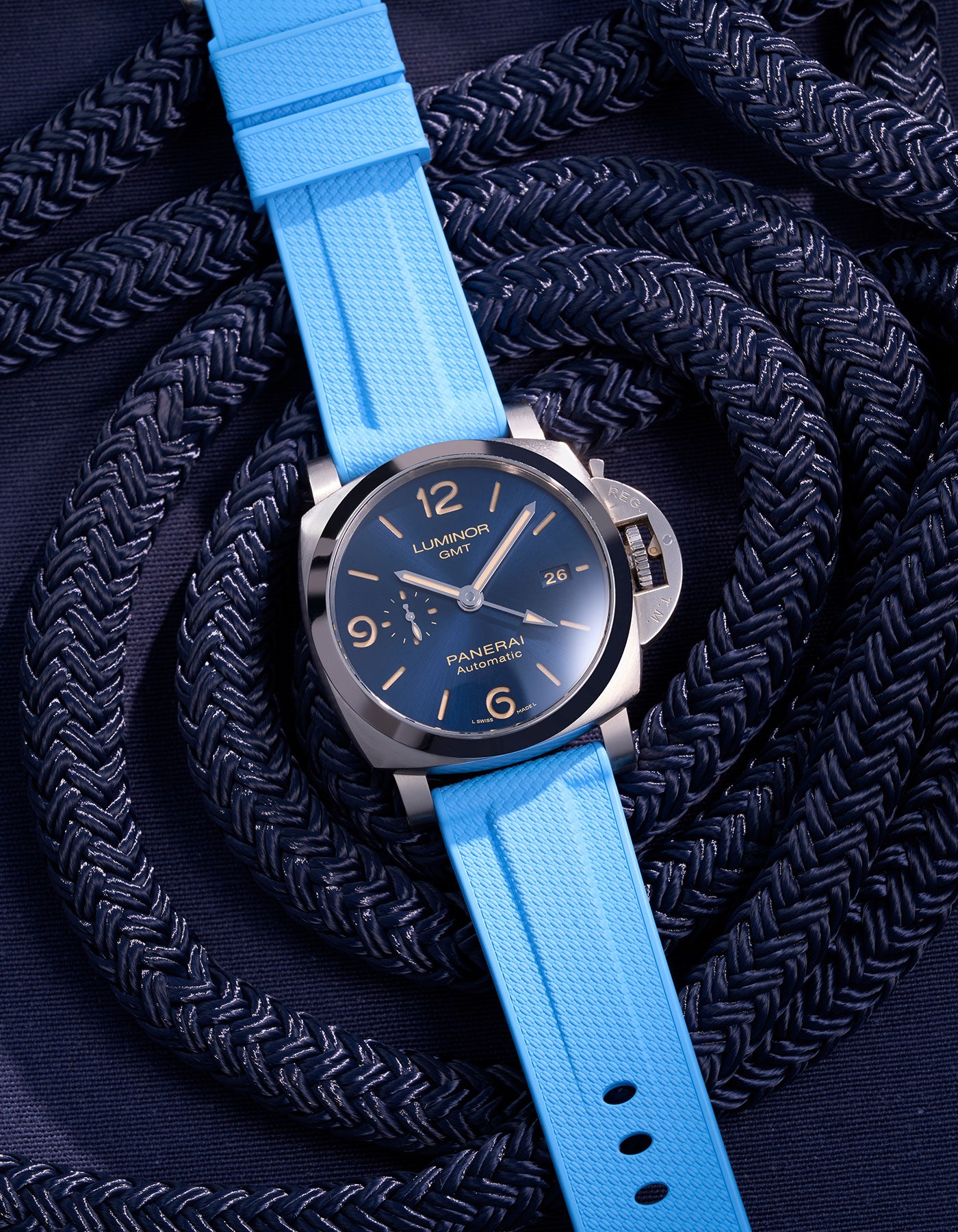 Panerai Luminor GMT Blue Dial with Light Blue Rubber Strap and Navy Rope Background – En kombination av italiensk elegans och schweizisk kvalitet i denna 44mm Panerai med ett påfallande ljusblått klockarmband, perfekt för aktiva livsstilar och nautiska miljöer. Klockans GMT-funktion och automatiska urverk gör den till en praktisk och stilren följeslagare, med det ljusblå bandet som framhäver klockans unika marina design.