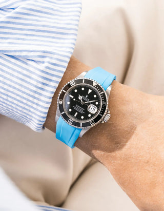 Capri Blue FKM rubber strap integrated for original clasp on Rolex Submariner 16610LN full steel, black ceramic bezel, black dial, straight front-facing wrist shot, smooth bright light blue strap texture, light blue striped linen shirt sleeve, cream background