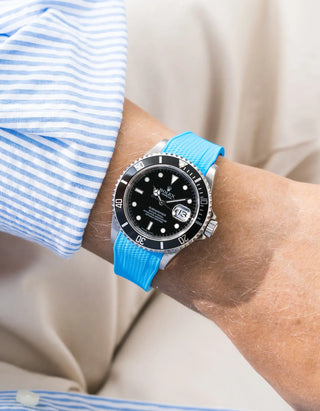Capri Blue Vertech FKM rubber strap integrated for original clasp on Rolex Submariner 16610LN full steel, black ceramic bezel, black dial, angled top-down wrist shot, Vertech crosshatch texture clearly visible on bright light blue strap, light blue striped linen shirt sleeve, cream background