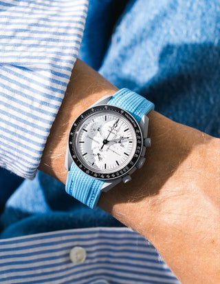 Capri blue rubber strap with fine diagonal ribbed texture on Omega x Swatch MoonSwatch Mission to the Moon 1965, wrist shot against blue denim jacket background, white dial with black subdials and black tachymeter bezel, white bioceramic case, blue and white striped shirt with white button visible at lower left