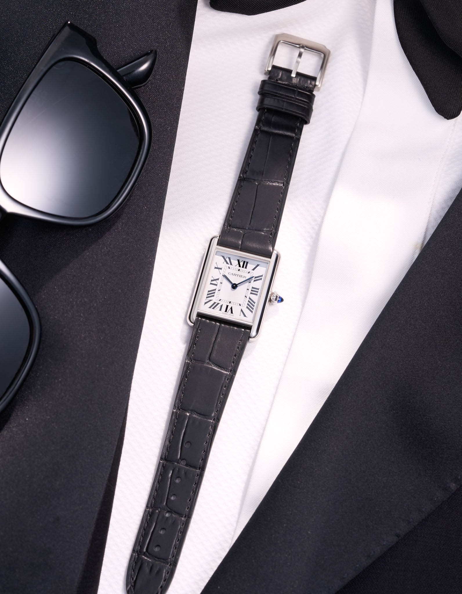 Cartier Tank Must Large with black embossed alligator leather strap, styled flat on white shirt and black suit with sunglasses beside