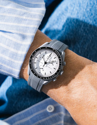 Dark grey rubber strap with fine cross-hatched ribbed texture on Omega x Swatch MoonSwatch Mission to the Moon 1965, wrist shot against blue fluffy textile background, white dial with black subdials and black tachymeter bezel, white bioceramic case, blue and white striped shirt with white button visible at lower right