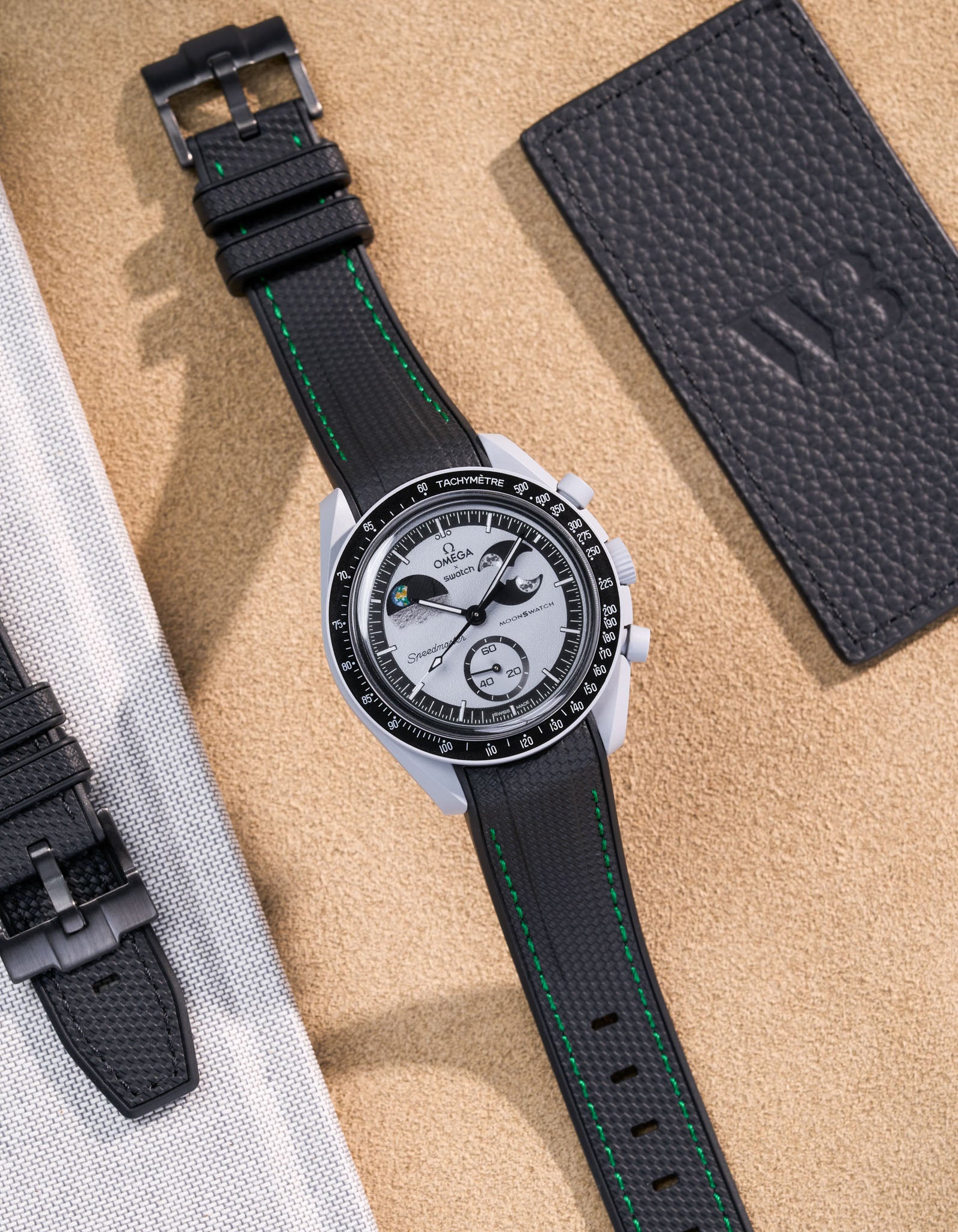 Flat lay of MoonSwatch Mission to EarthPhase with black rubber strap with green stitches, watch facing up with clasp visible