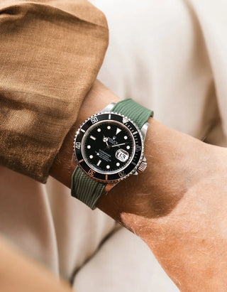 Green Vertech FKM rubber strap integrated for original clasp on Rolex Submariner 16610LN full steel, black ceramic bezel, black dial, angled wrist shot from left side, Vertech crosshatch texture clearly visible on olive green strap, brown linen jacket sleeve, cream background