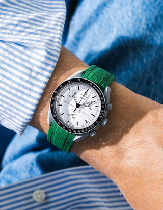 Bright green rubber strap with diagonal ribbed texture on Omega x Swatch MoonSwatch Mission to the Moon 1965, wrist shot against blue denim jacket background, white dial with black subdials and black tachymeter bezel, white bioceramic case, blue and white striped shirt sleeve visible at left