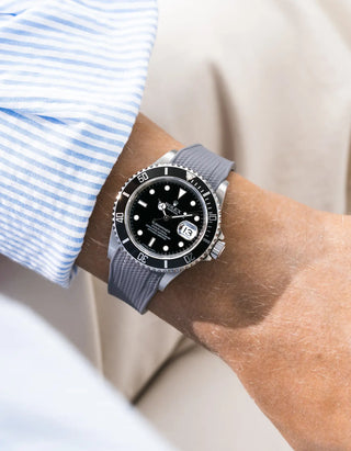 Grey Vertech FKM rubber strap integrated for original clasp on Rolex Submariner 16610LN full steel, black ceramic bezel, black dial, angled wrist shot from left side, Vertech crosshatch texture visible on strap, blue striped shirt cuff, white and beige background