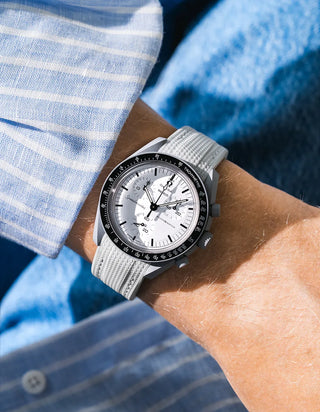 Grey rubber strap with fine cross-hatched ribbed texture on Omega x Swatch MoonSwatch Mission to the Moon 1965, wrist shot against blue fluffy textile and blue and white striped shirt background, white dial with black subdials and black tachymeter bezel, white bioceramic case, white button on shirt visible at lower left