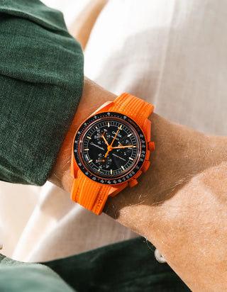 Lava orange rubber strap with vertical ribbed texture on Omega x Swatch MoonSwatch Mission on Earth Lava, wrist shot against cream fabric background, black dial with orange chronograph hands, dark green jacket visible at upper and lower left