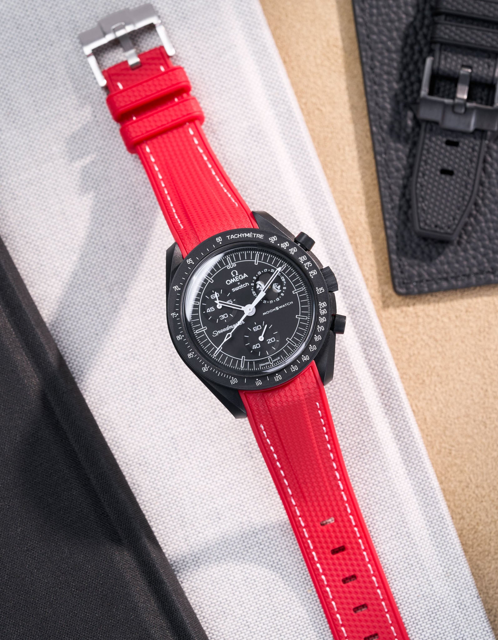 MoonSwatch Mission to Moonphase new Moon with vertech red rubber strap with white stitching placed on white fabric surface next to black pouch