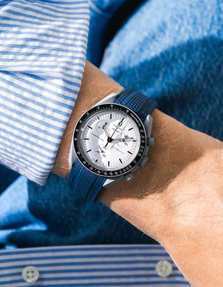 Navy blue rubber strap with diagonal ribbed texture on Omega x Swatch MoonSwatch Mission to the Moon 1965, wrist shot against blue denim jacket background, white dial with black subdials and black tachymeter bezel, white bioceramic case, blue and white striped shirt with white buttons visible at left and lower right