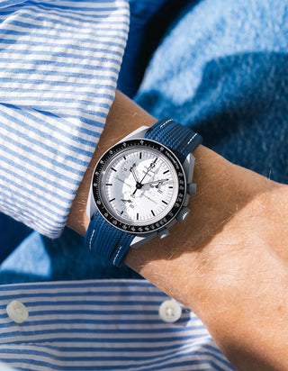 Navy blue rubber strap with white contrast stitching and diagonal ribbed texture on Omega x Swatch MoonSwatch Mission to the Moon 1965, wrist shot against blue denim jacket background, white dial with black subdials and black tachymeter bezel, white bioceramic case, blue and white striped shirt with three white buttons visible at lower left