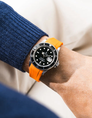 Orange FKM rubber strap integrated for original clasp on Rolex Submariner 16610LN full steel, black ceramic bezel, black dial, angled wrist shot from left side, smooth orange strap, navy blue knit sweater sleeve, beige background