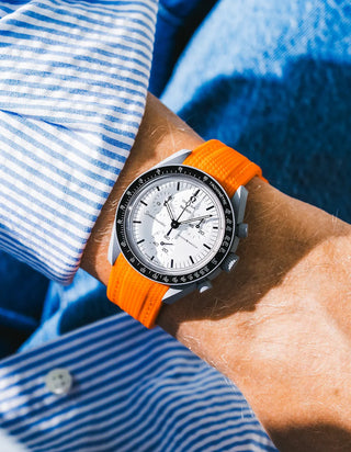 Bright orange rubber strap with fine diagonal ribbed texture on Omega x Swatch MoonSwatch Mission to the Moon 1965, wrist shot against vivid blue background, white dial with black subdials and black tachymeter bezel, white bioceramic case, blue and white striped shirt with white button visible at lower left, shot in direct sunlight