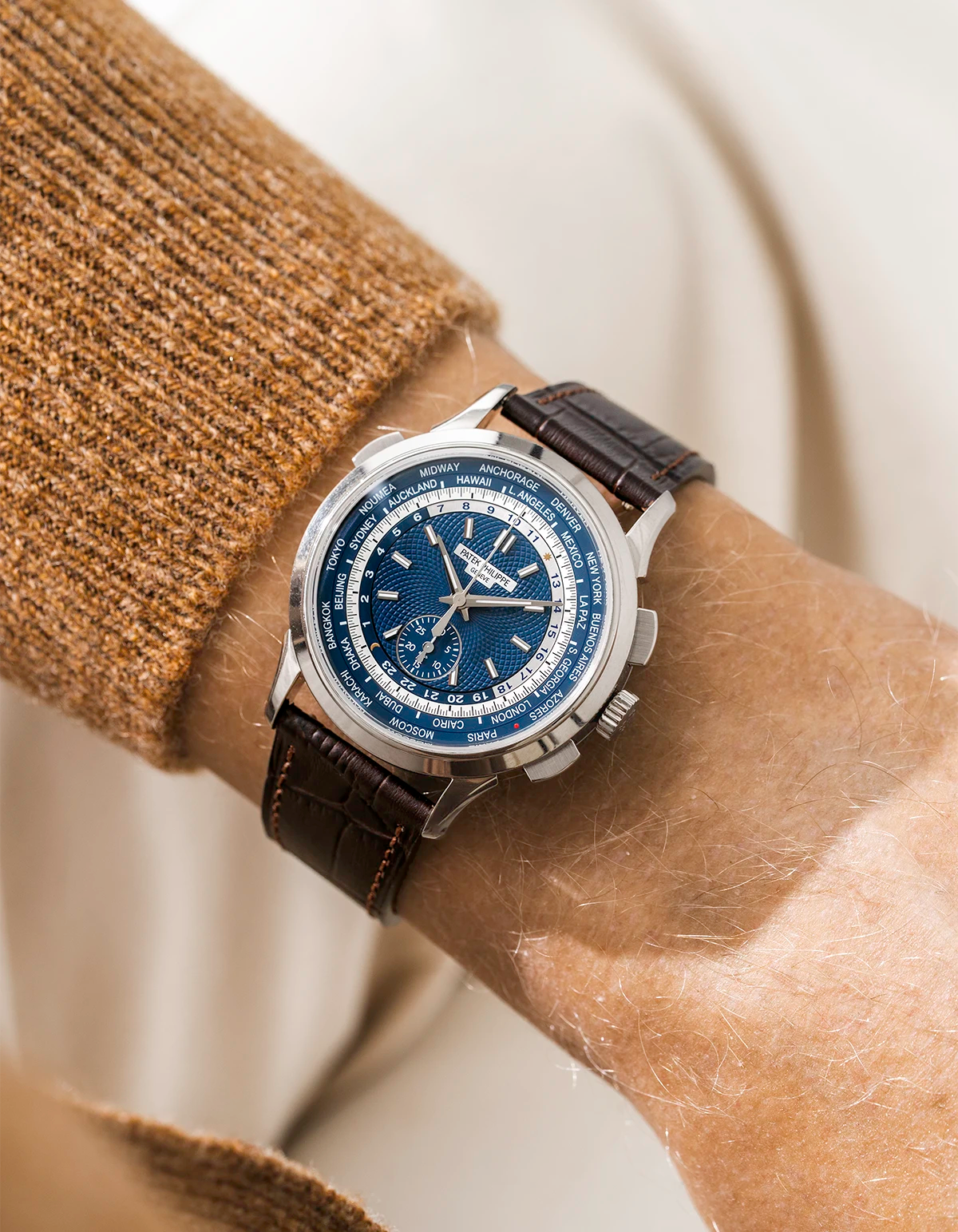 Patek Worldtimer watch with a blue face and dark brown wristbuddys alligator watch strap on a wrist, against a neutral background
