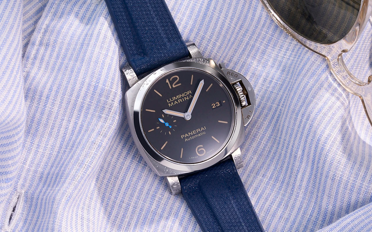 Panerai Luminor Marina with navy blue rubber strap resting on a light blue striped shirt next to sunglasses